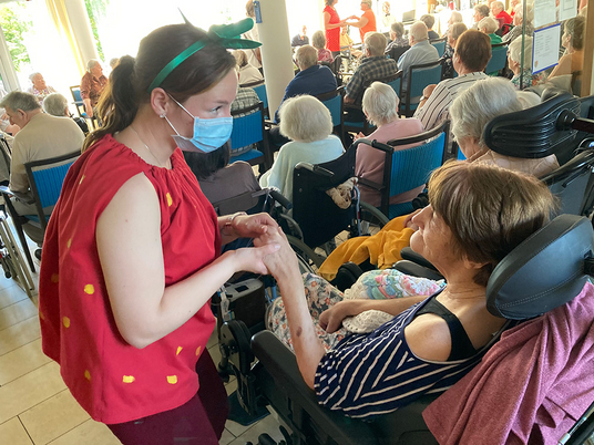 Eine Mitarbeiterin hält die Hand einer Bewohnerin im Rollstuhl im vollen Saal | Immanuel Haus am Kalksee | Seniorenzentrum | Erdbeerfest